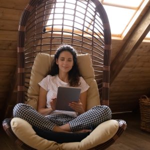 Happy focused Latin woman relaxing in comfortable armchair in country house, using virtual app on tablet, working or studying from home, watching learning webinar, making video call, reading book